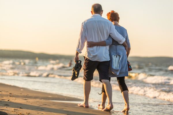 Ein älteres Paar geht barfuß und eng umschlungen bei Sonnenuntergang am Strand entlang.