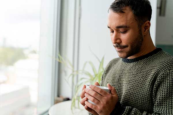 Mann steht mit einer Tasse in beiden Händen am Fenster und blickt nach unten.