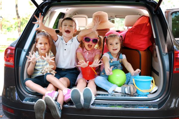 Vier Kinder sitzen zwischen Reisegepäck im offenen Kofferraum eines Autos und strecken fröhlich die Hände nach vorn.