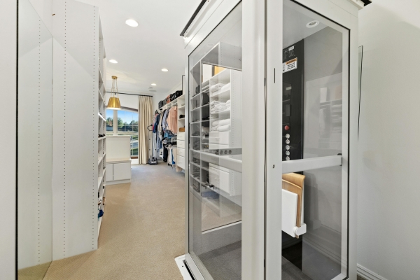 Verglaster Senkrechtlift mit Bedienelementen befindet sich in einem hellen, begehbaren Kleiderschrank mit Regalen und Kleidung.
