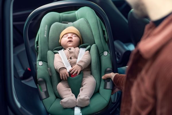 Baby liegt angeschnallt in einer Babyschale im Auto, während eine Person danebensteht.