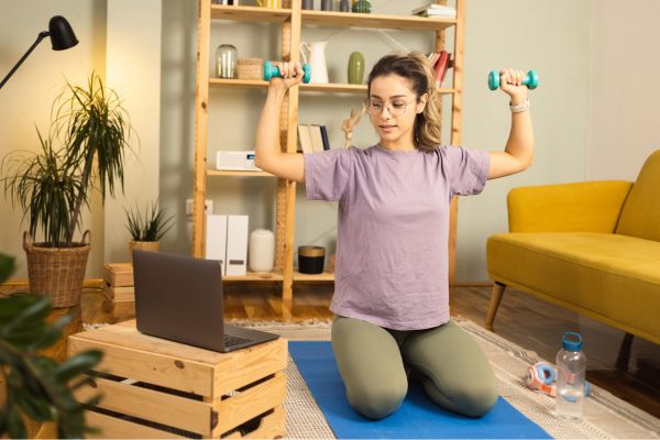 Junge Frau kniet auf einer blauen Yogamatte im Wohnzimmer und führt mit kleinen Hanteln gezielte Schulter- und Rückenübungen durch; vor ihr steht ein Laptop, im Raum befinden sich Sofa, Regal, Pflanzen und Trainingszubehör.
