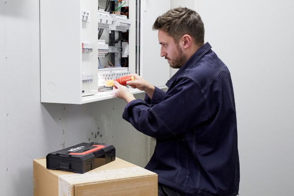 Handwerker arbeitet mit einem Schraubendreher an einem geöffneten Elektroschaltkasten, neben ihm steht ein Werkzeugkoffer.