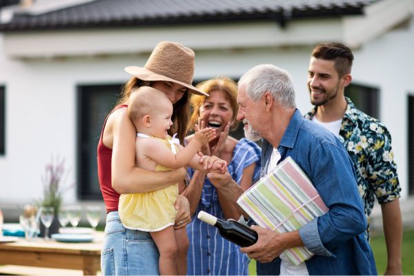 Mehrgenerationenfamilie feiert zusammen im Garten, ein älterer Mann hält ein Geschenk und eine Weinflasche.