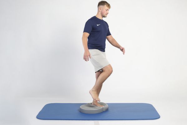 Mann steht auf einem Balancekissen auf einem Bein und trainiert das Gleichgewicht auf einer Matte.
