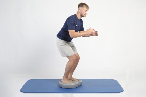 Mann macht eine Kniebeuge auf einem Balancekissen und trainiert das Gleichgewicht auf einer Matte.