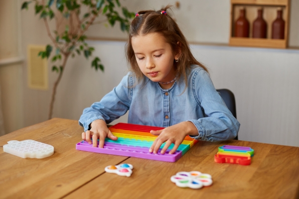 Mädchen sitzt am Tisch und beschäftigt ihre Hände mit einem bunten Fidget-Spielzeug.