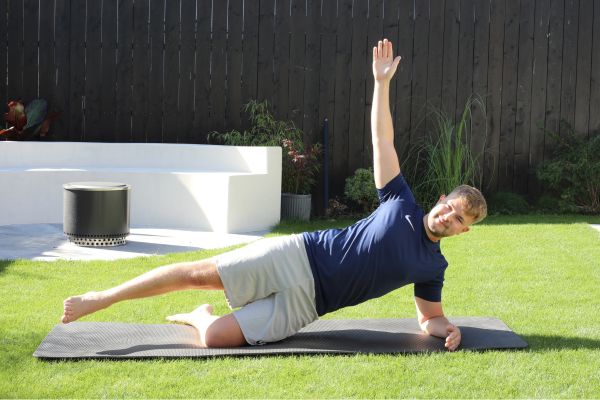 Ein Mann liegt auf einer Matte auf einem Rasen und führt eine Dehnübung durch.