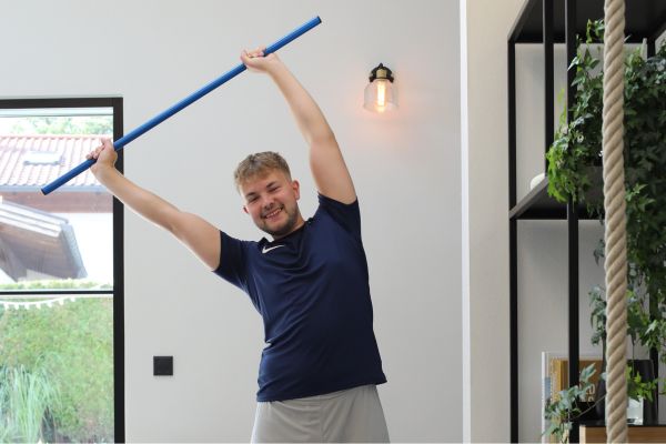 Ein Mann führt i einem Zimmer eine Seitwärtsbewegung mit einem Turnstab aus. Link führt zu einer Seite mit Rückenübungen mit Turnstab.