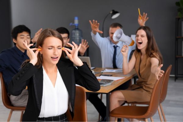 Frau hält sich im Büro die Ohren zu, während Kollegen im Hintergrund lautstark durcheinanderreden.