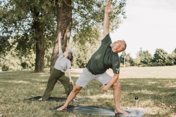 Zwei ältere Menschen machen auf Gymnastikmatten im Park eine seitliche Dehnübung mit ausgestrecktem Arm.