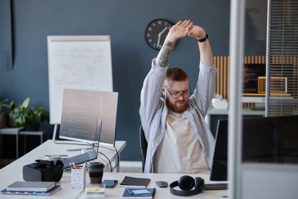 Mann sitzt am Schreibtisch im Büro und streckt beide Arme über den Kopf.