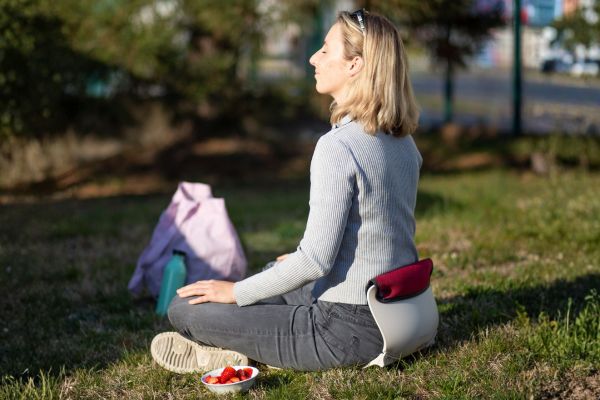 Frau sitzt auf einer Sitzauflage im Park und meditiert entspannt im Schneidersitz auf einer Wiese.
