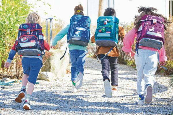 Vier Kinder laufen mit Schulranzen auf dem Rücken gemeinsam einen Kiesweg entlang.