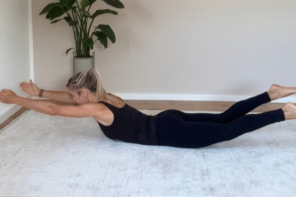Frau führt auf einer Matte die Yoga-Übung Heuschrecke in Bauchlage aus und hebt Arme, Oberkörper und Beine an.