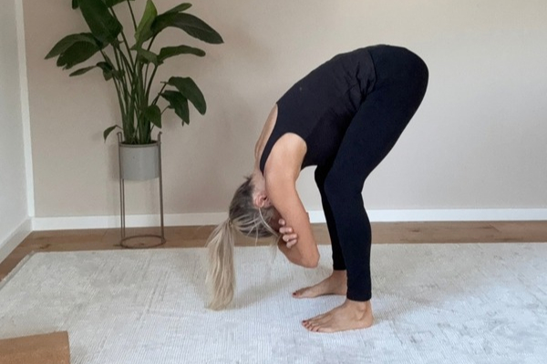 Frau führt auf einer Matte eine stehende Yoga-Vorbeuge mit leicht gebeugten Knien und nach unten hängendem Oberkörper aus.