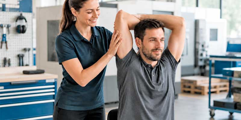 Physiotherapeutin unterstützt einen Mitarbeiter in Arbeitskleidung bei einer Schulter- und Armdehnung in einer Werkstatt.