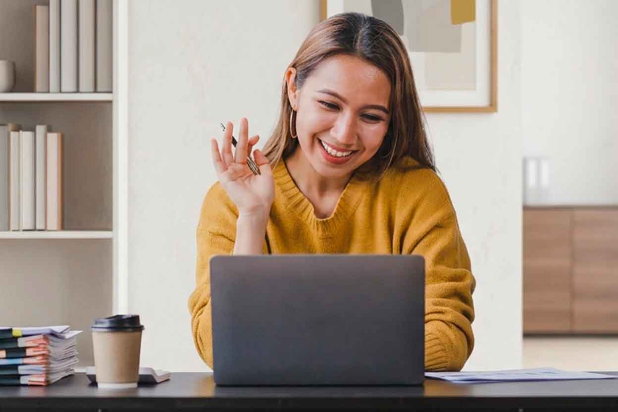 Frau sitzt am Schreibtisch vor Laptop, lächelt und nimmt an einem Online-Seminar teil, Unterlagen und Kaffee daneben