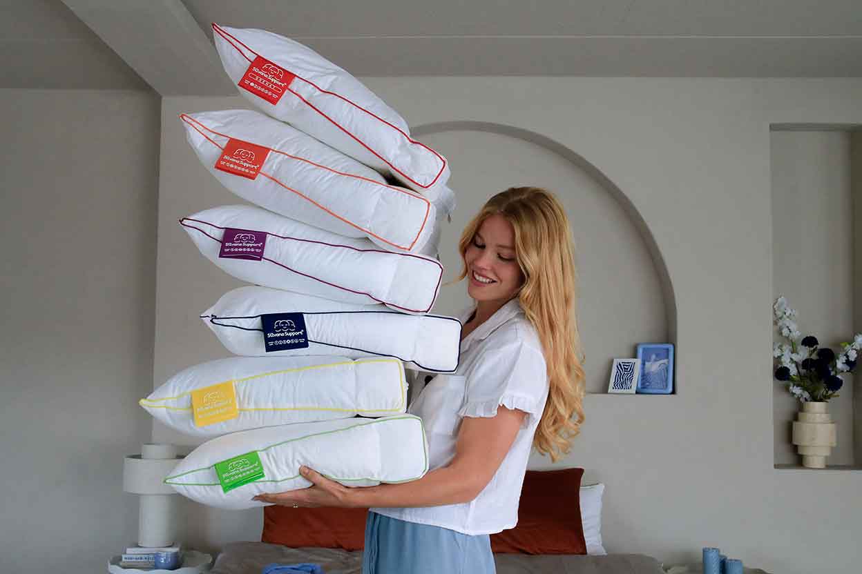 Frau trägt mehrere gestapelte Kopfkissen in einem hellen Schlafzimmer.