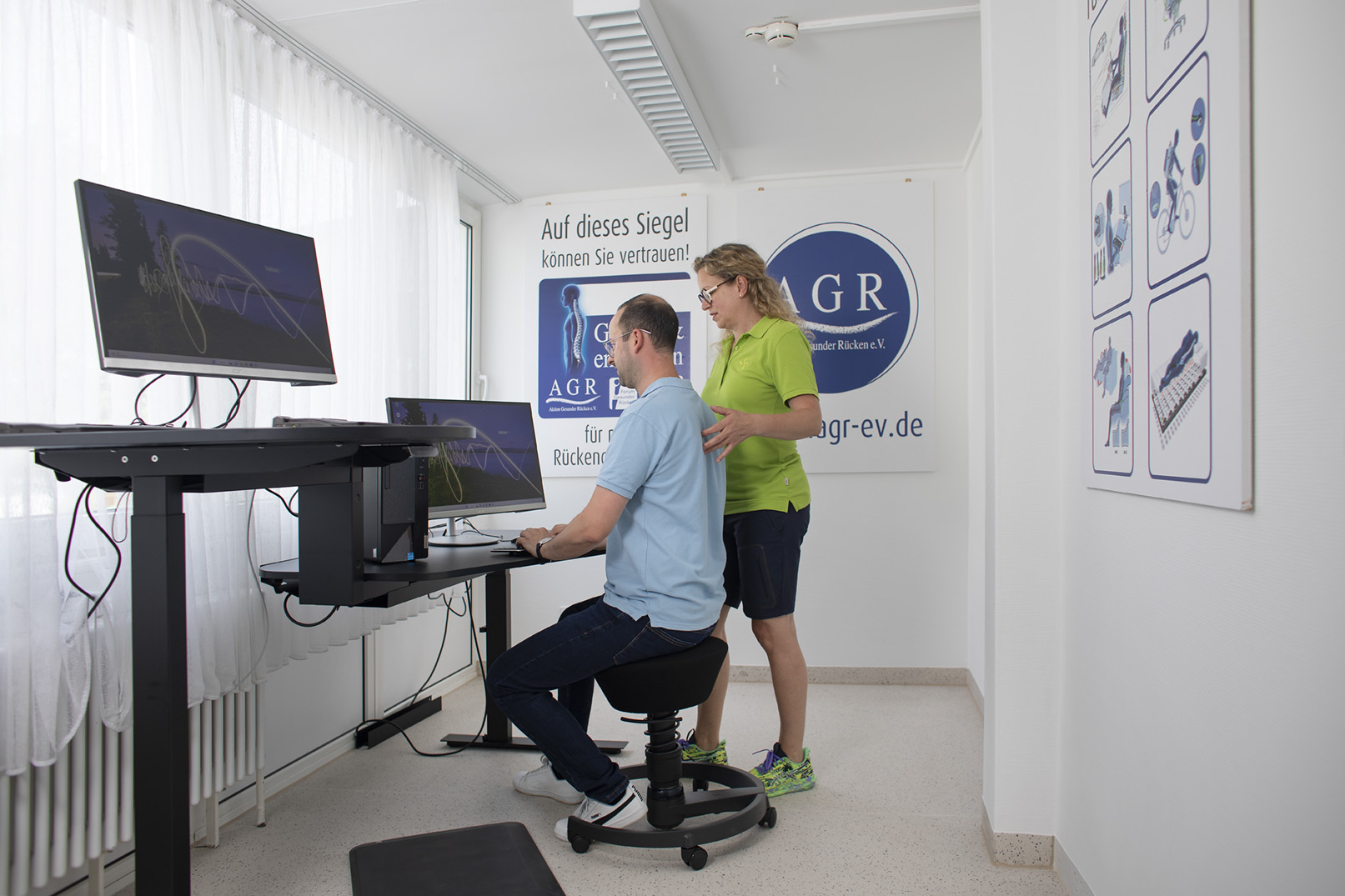 Frau berät Mann beim ergonomischen Arbeiten im Sitzen und Stehen am Schreibtisch