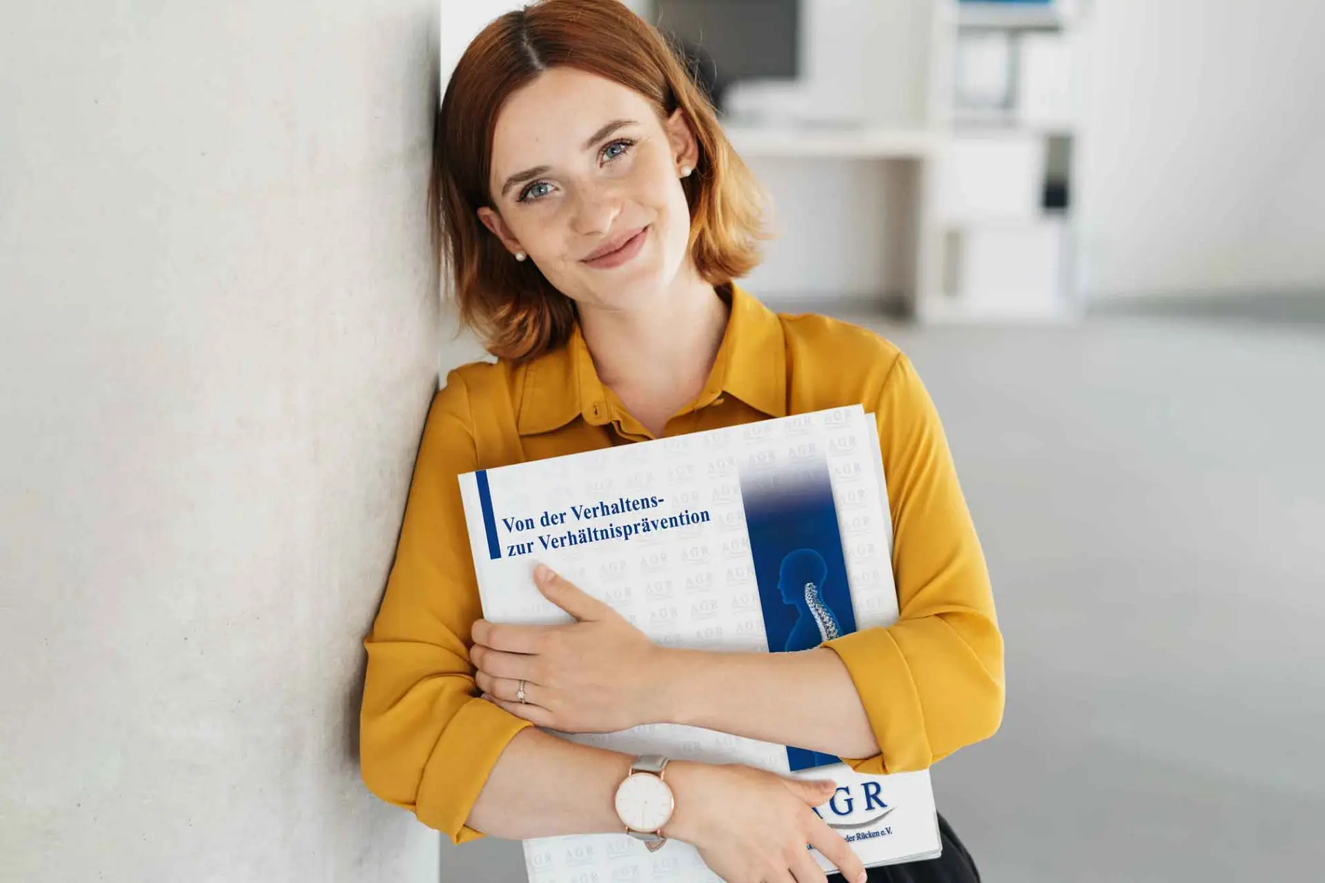 Informationen zum AGR- Fernlehrgang "Von der Verhaltens- zur Verhältnisprävention" Junge Frau lehnt mit Schulter an einer Wand und hält einen Ordner im Arm. Führt zur Infoseite Fernlehrgang