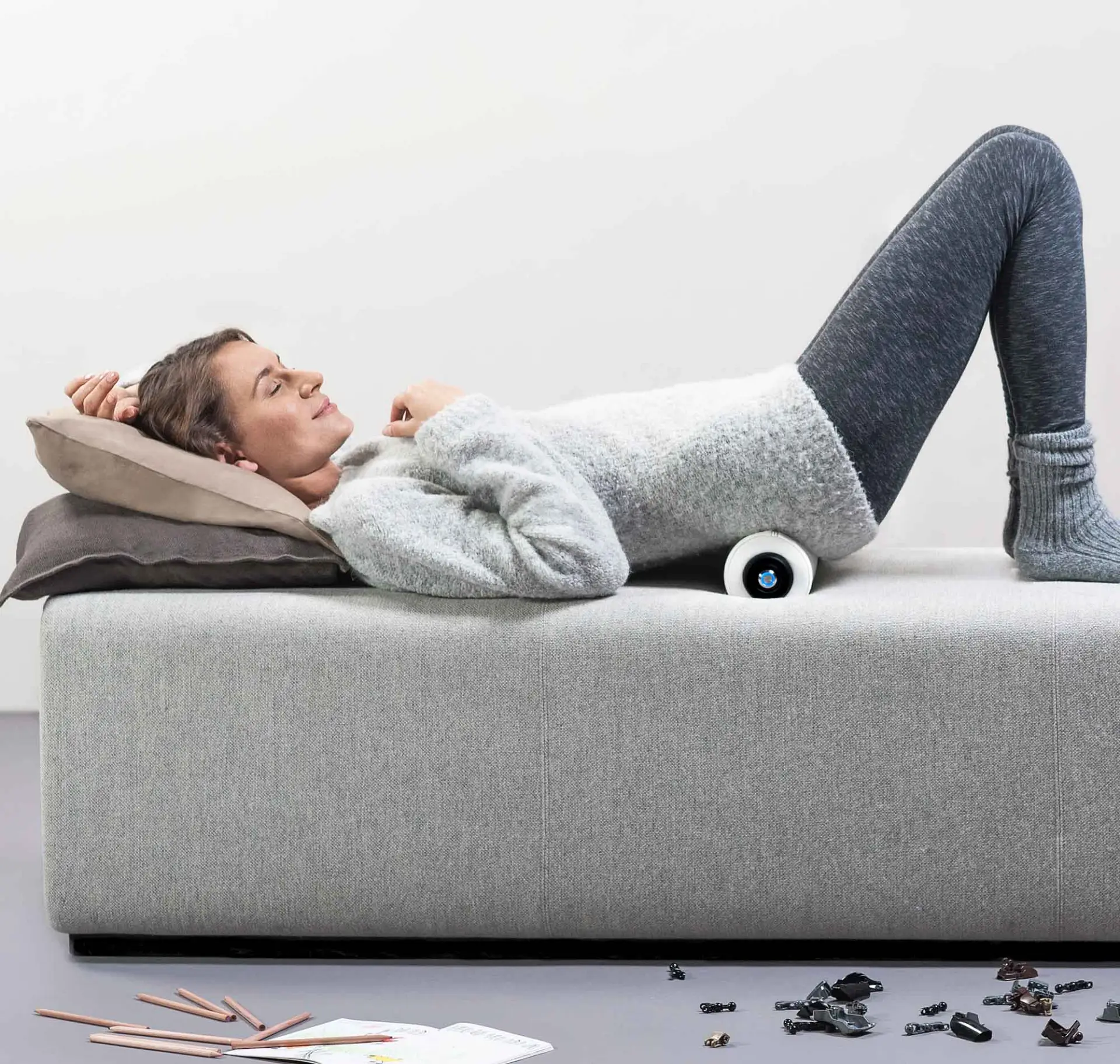 Eine Frau liegt auf einem Sofa mit einer Massagerolle im unteren Rücken, sie entspannt sich dabei.