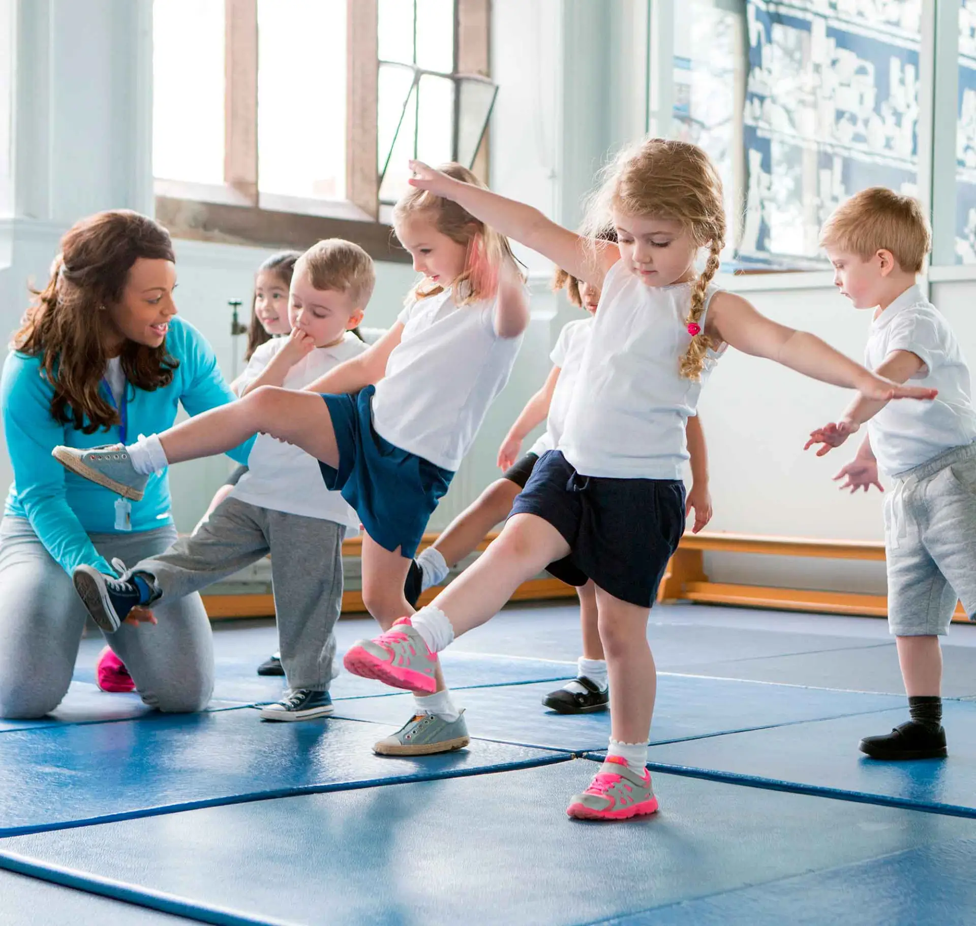 Ein paar kleine Kinder turnen in einer Turnhalle, eine Frau hilft einen Jungen bei den Übungen.