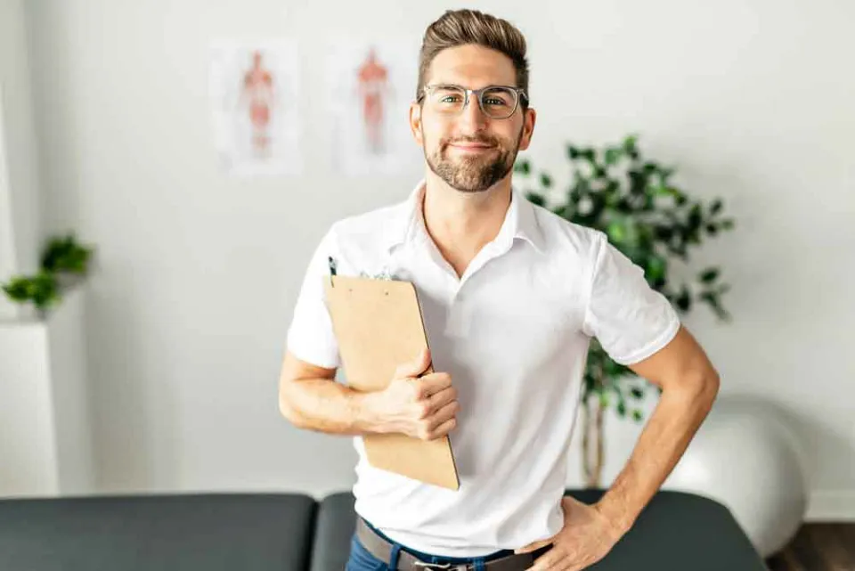 Freundlicher Mann in weißem Polo-Shirt hält Klemmbrett und steht vor Anatomie-Postern in einer Praxis